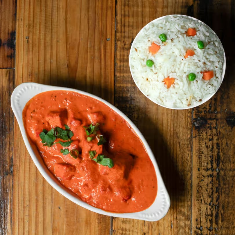 Indian butter chicken in a white dish with cilantro, and a bowl of rice with peas and carrots.